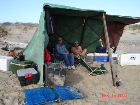 1995 Ford Bronco - Two men relax under a makeshift tarp shelter on a sandy beach, surrounded by coolers and camping gear.