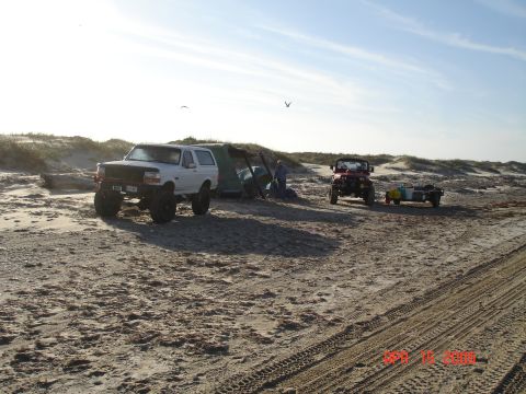1995 Ford Bronco - Two off-road vehicles are parked on a sandy beach near a tent under a partly cloudy sky.