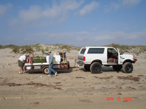 1995 Ford Bronco - A white 1995 Ford Bronco is parked on a sandy beach, towing a trailer loaded with gear.