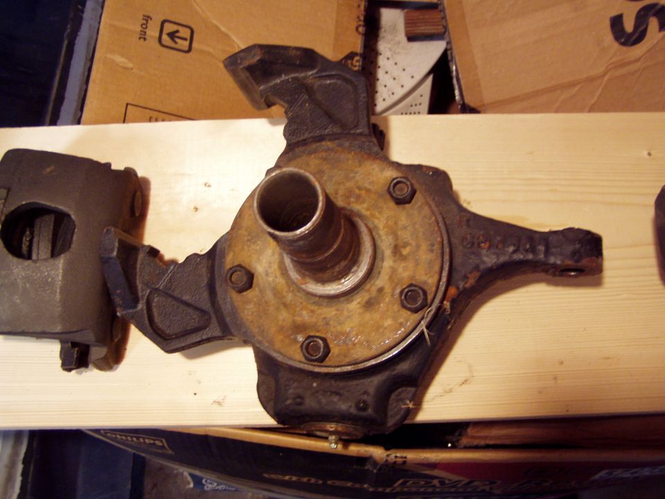 Vehicle axle assembly and brake caliper on a wooden surface. The axle has a rusty hub and is bolted to a bracket