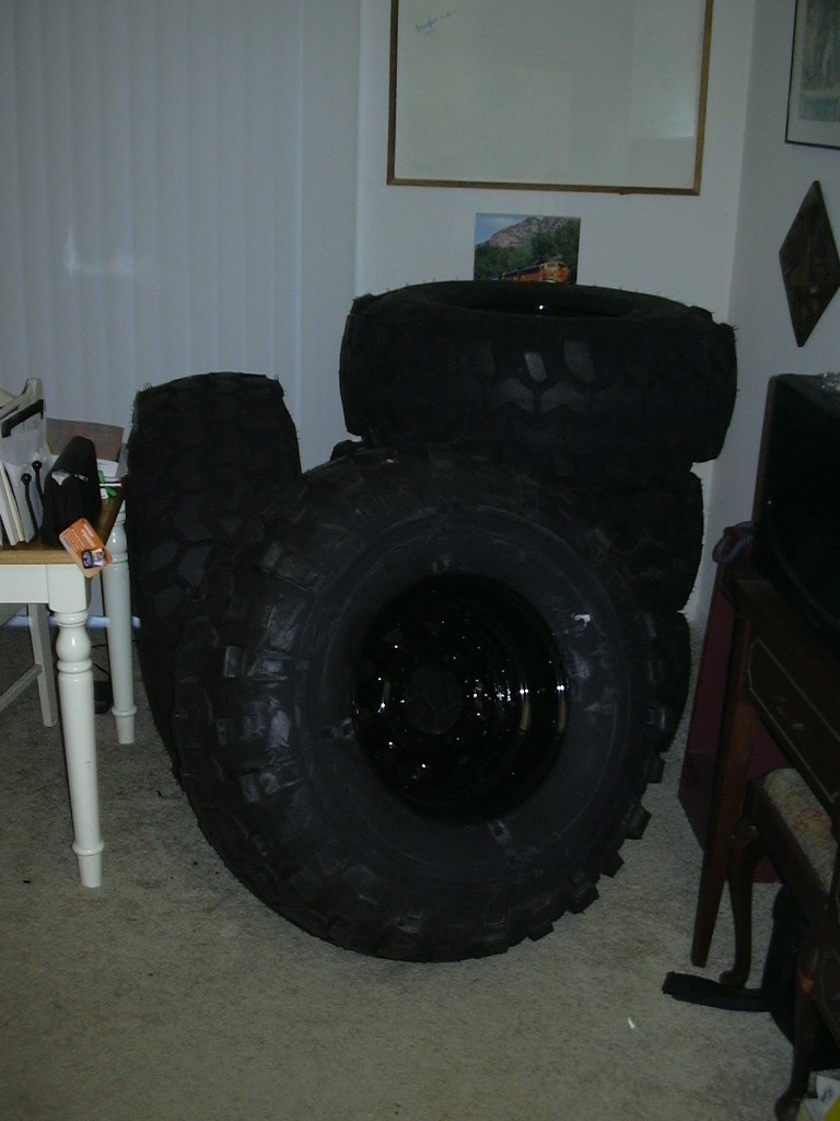 Several large off-road tires stacked indoors, one in foreground is black with a shiny rim