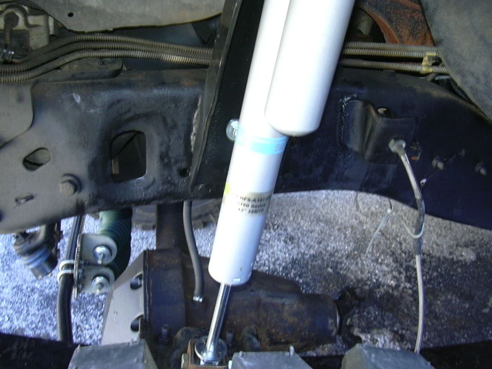 Close-up of a white Bilstein shock absorber attached to a black metal frame with visible brake lines and suspension components