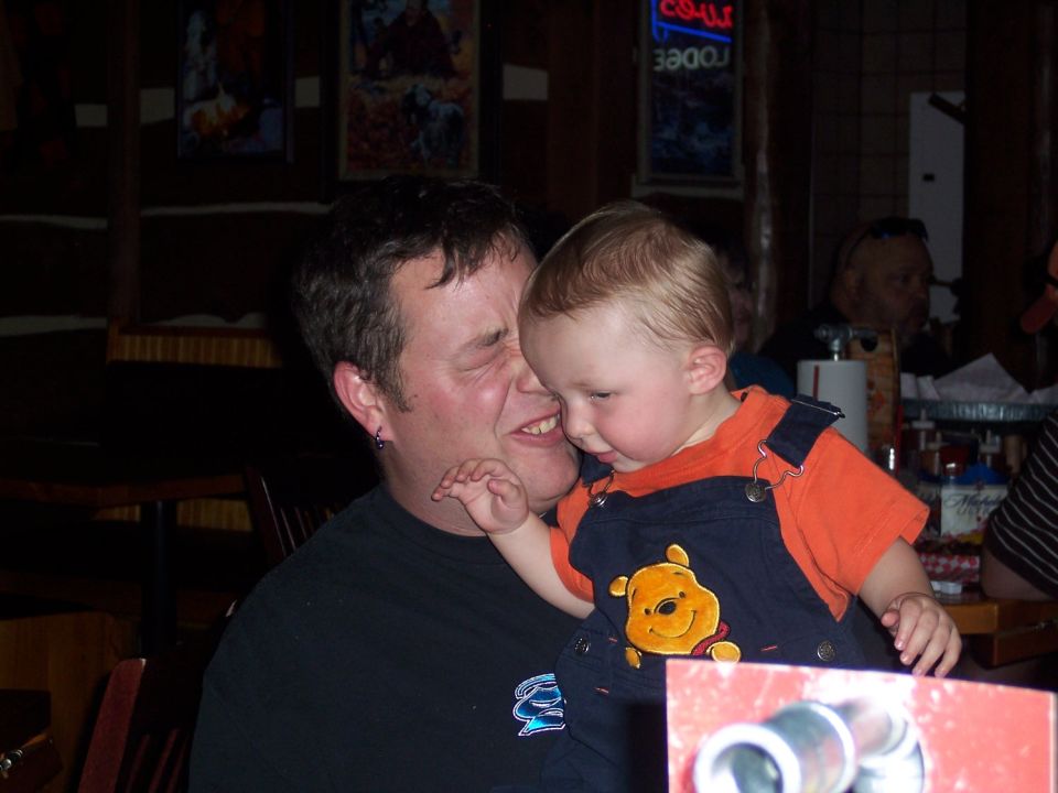 Man with a black t-shirt holding a baby wearing orange shirt and blue overalls with Winnie the Pooh