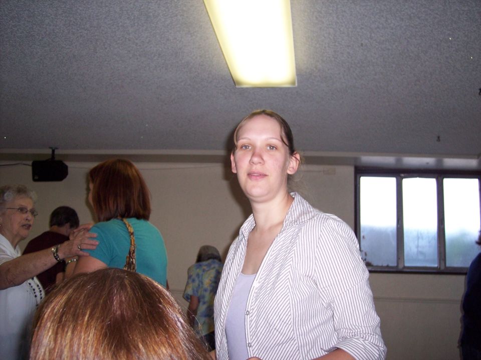 Woman in a striped button-up shirt and tank top stands indoors with other people blurred in the background
