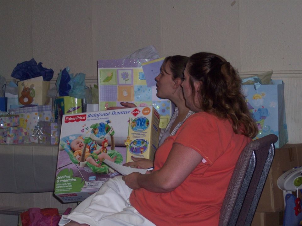 Two women look at a Fisher-Price Rainforest Bouncer box at a baby shower. Gifts are visible in the background