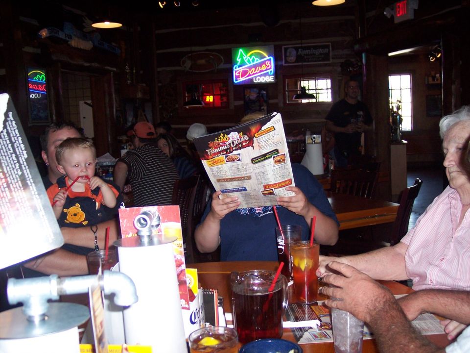 People sit at a table in a log cabin-style restaurant, looking at menus. A baby in a Winnie the Pooh outfit is held by someone