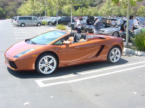 1993 Ford Probe - An orange convertible sports car is parked in a lot alongside other vehicles, with people gathered around some of them.