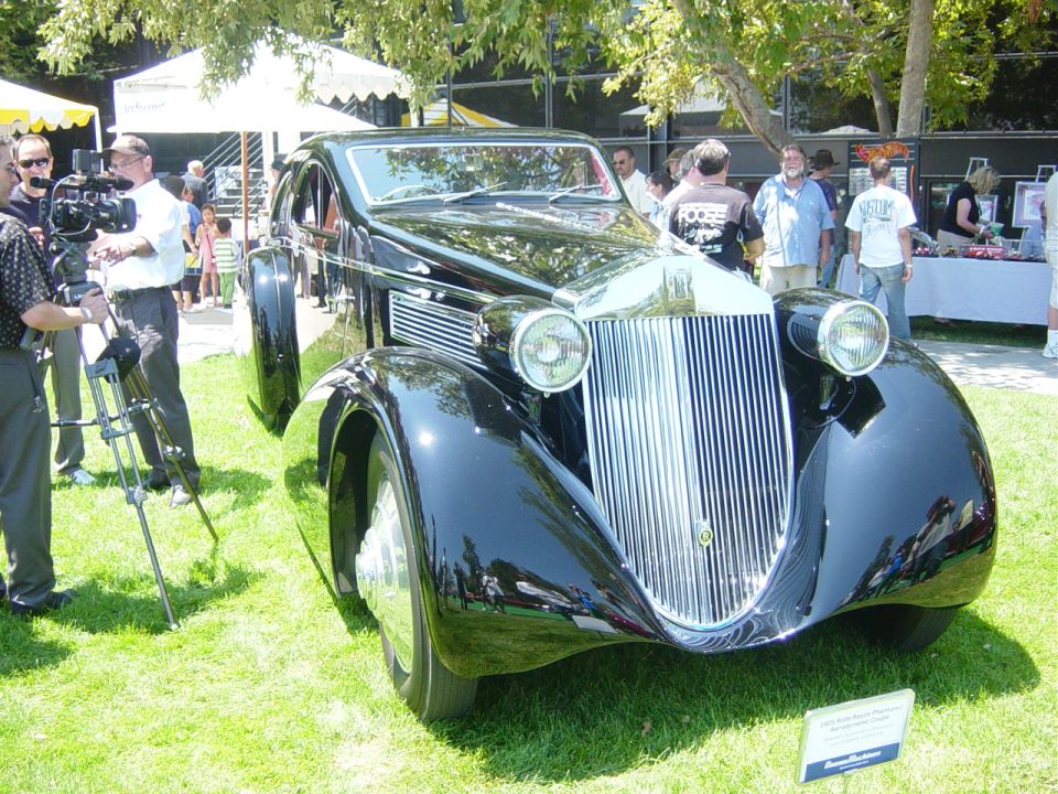Black vintage car with chrome grille and headlights on grass, camera crew filming