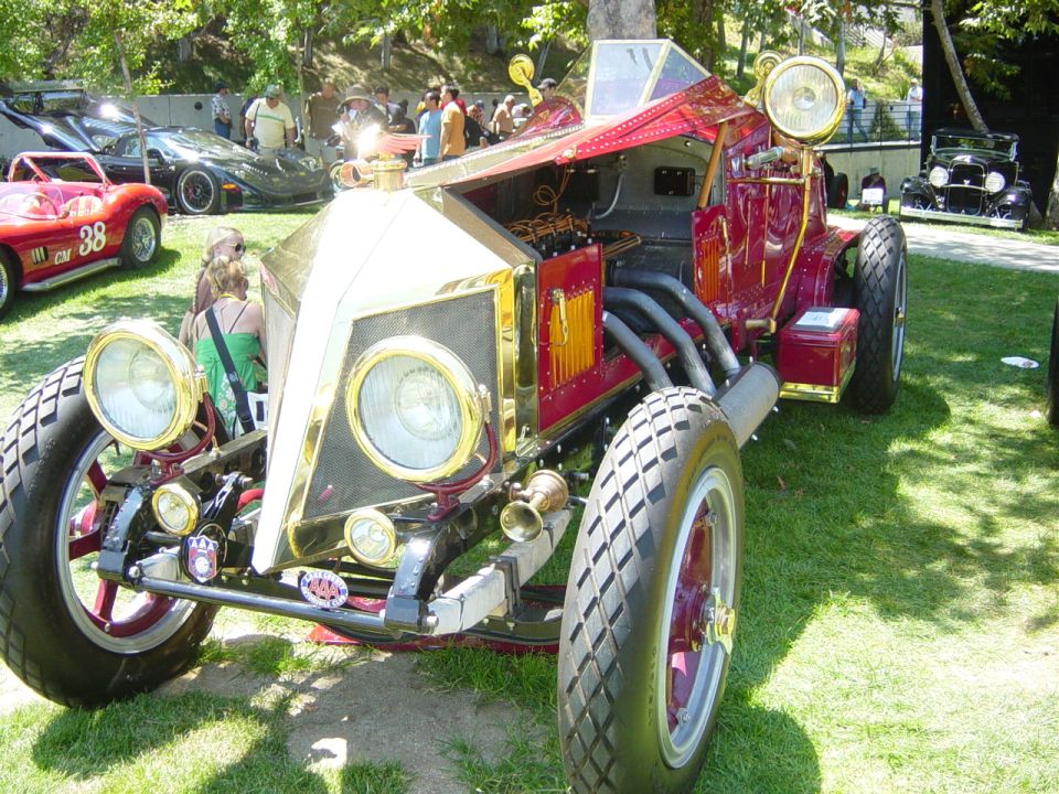 Front view of a custom hot rod with a gold grille, exposed engine, and red bodywork. Other cars are visible in the background