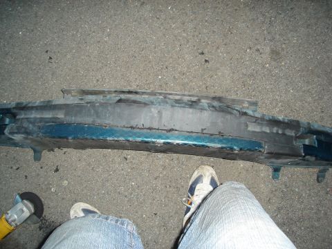 1995 Mazda MX-6 - Car bumper part lying on asphalt next to a grinder and a person's legs.