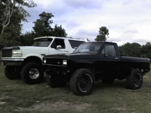 1995 Ford Bronco - A white 1995 Ford Bronco is parked next to a black pickup truck under a cloudy sky.