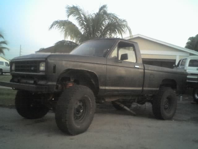 Lifted pickup truck with aftermarket bumper and large tires. Another white vehicle is parked behind it