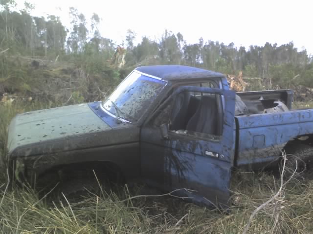 Muddy blue pickup truck with door open, stuck in tall grass and brush