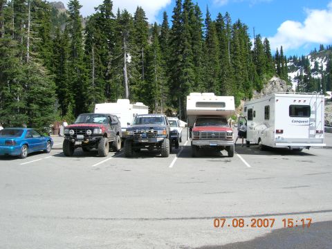 1996 Ford Bronco - Several vehicles, including modified SUVs and RVs, are parked in a lot with a scenic mountain and forest view. The photo was taken on July…