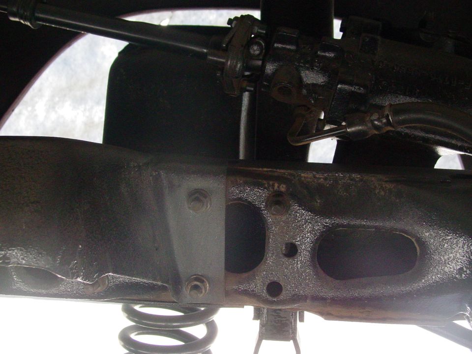 Close-up of vehicle frame with suspension spring visible. Steering linkage is also in view