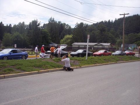 1995 Ford Probe - A group of cars, including a blue Ford Probe, are gathered with people on a sunny day, while a photographer captures the scene.