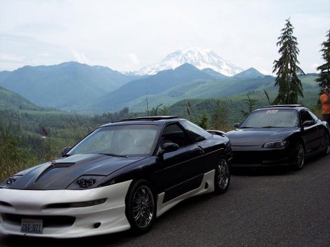 1995 Ford Probe - Two modified coupes are parked on a scenic mountain road with a snow-capped peak in the background.