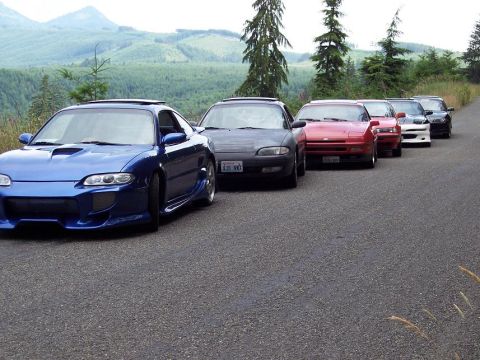 1995 Ford Probe - A line of sports cars drives down a road with a wooded hillside in the background.