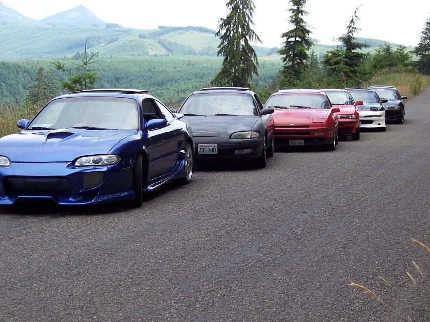 Blue sports car with aftermarket body kit, followed by four other cars in a line on a paved road