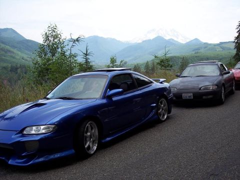1995 Ford Probe - Two coupes drive on a scenic mountain road with a range of mountains in the background.