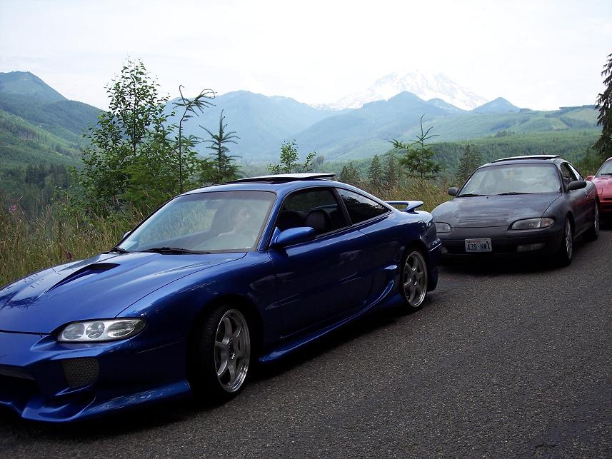 Blue coupe with aftermarket body kit and spoiler, parked behind a dark sedan on a mountain road. Mountains and trees in background