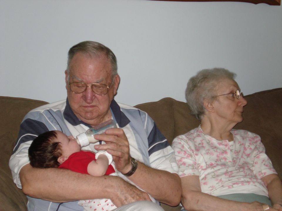 Grandfather feeding a baby with a bottle while a grandmother sits nearby