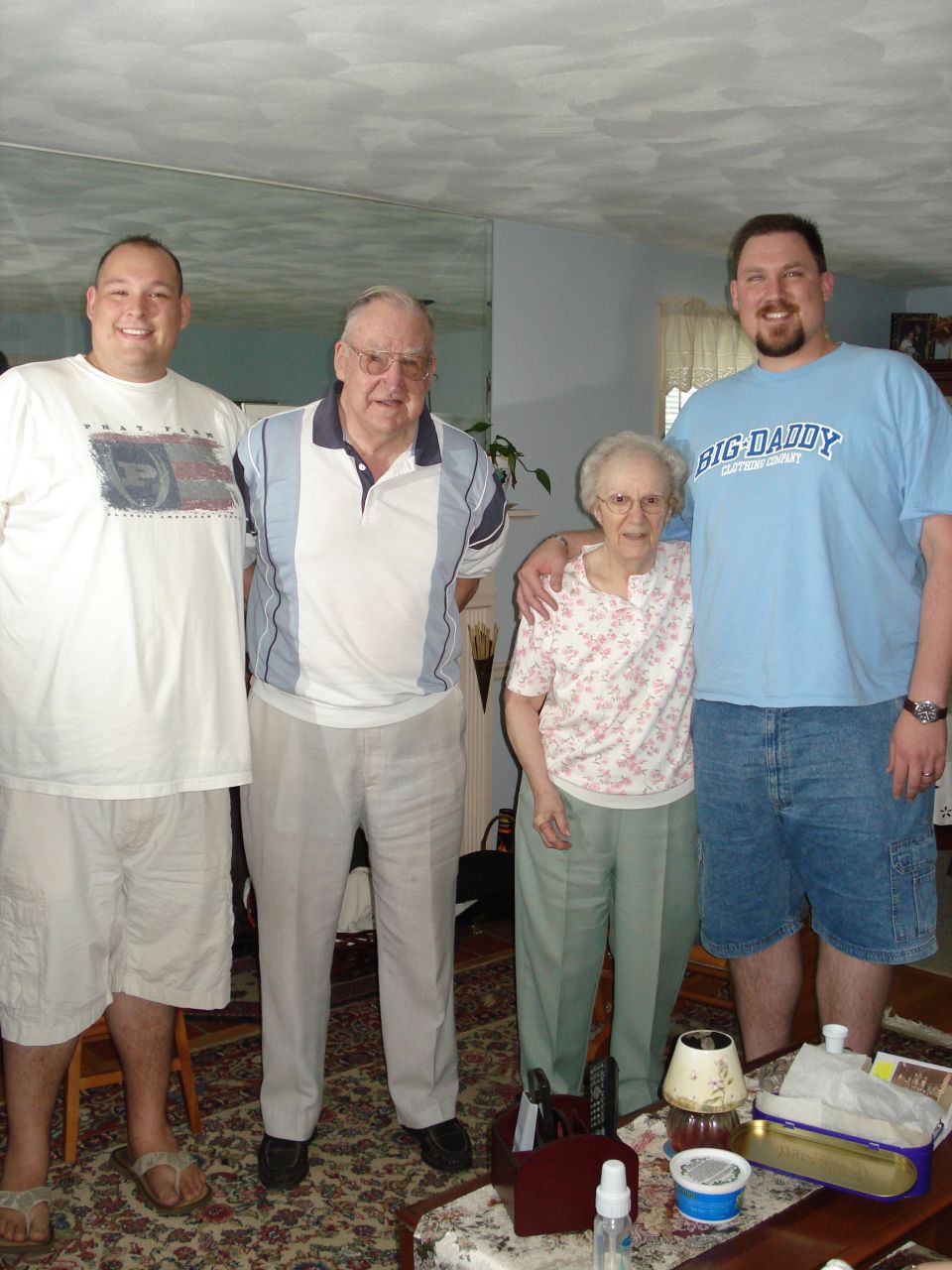 Four people, two men, one woman, and one older man, stand together indoors. The man on the far right wears a blue t-shirt with "BIG DADDY" printed on it