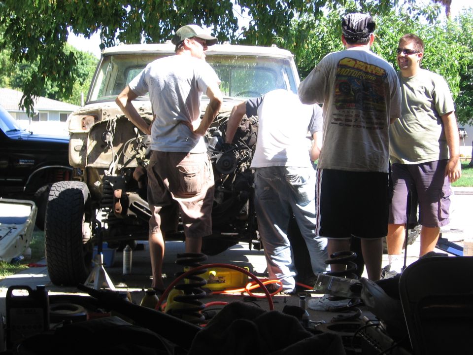 Ford F-series truck, front end disassembled, with three men standing around it. Another dark SUV is parked to the left