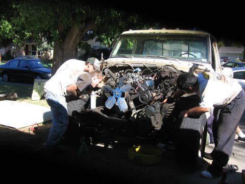1989 Ford Bronco - Mechanics are gathered around the open engine bay of a truck, appearing to be in the middle of repairs or maintenance.