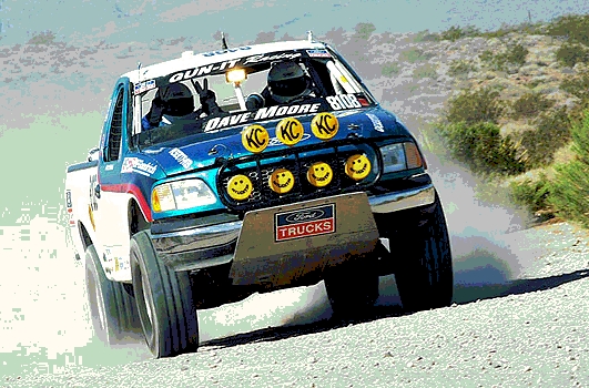 Blue and white 2000 Ford F-150 racing truck with KC lights and smiley faces on the grille, kicking up dust on a dirt road