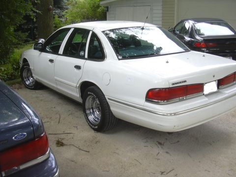1997 Ford Crown Victoria - A white 1997 Ford Crown Victoria is seen from a three-quarter front view, aftermarket chrome wheels and parked alongside other vehicles.