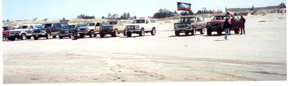 Long row of parked 4x4 trucks on a beach with a Ford-logo banner trailer at right