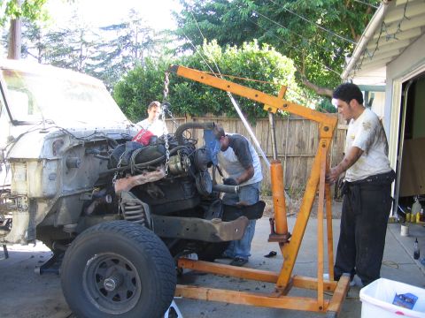 1989 Ford Bronco - Workers use an engine hoist to remove the engine from a disassembled truck, suggesting a repair or restoration project.