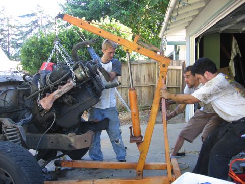 1989 Ford Bronco - Workers use an engine hoist to remove a large engine from a vehicle's chassis in a driveway.