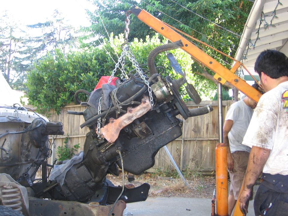 V8 engine and transmission hanging from an orange engine hoist during a driveway pull