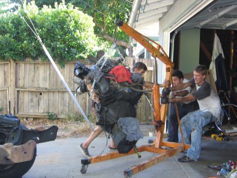 1989 Ford Bronco - Three men maneuver an engine being lifted by an engine hoist outside a garage.
