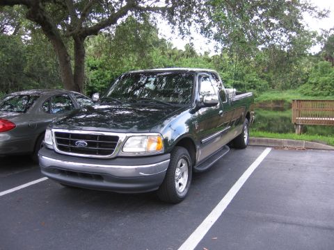 2001 Ford F-150 - A dark green Ford F-150 pickup truck is parked in a lot with a pond and wooden bench in the background.