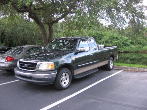2001 Ford F-150 - A dark green 2001 Ford F-150 pickup truck is parked in a lot beside a silver sedan, with trees and water in the background.