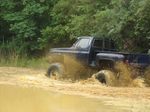 1976 Chevrolet Blazer - A dark blue pickup truck navigates a muddy terrain, kicking up water and dirt.