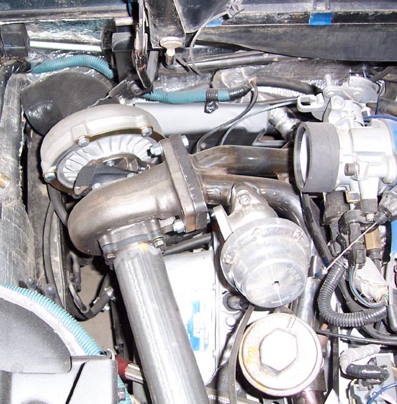 Close-up of a turbocharger, exhaust manifold, and engine plumbing. SuperMotors.net logo visible