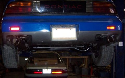 1988 Pontiac Fiero - The rear of a blue Pontiac Fiero GT is shown on a lift, with another vehicle positioned underneath it.