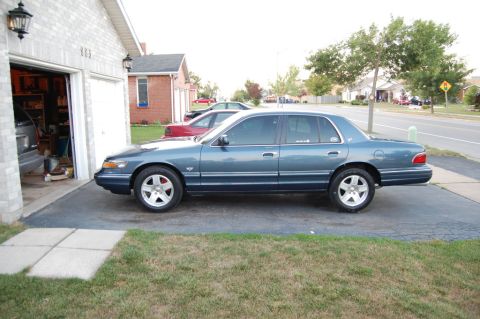 1985 Mercury Grand Marquis - A blue sedan is parked in a driveway next to a garage, with another red sedan visible behind it.