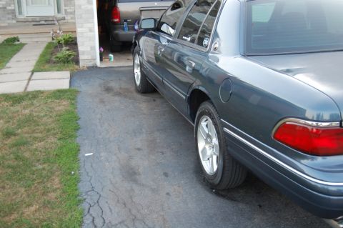 1985 Mercury Grand Marquis - The rear quarter panel and wheel of a dark blue sedan are visible, with a gray SUV parked in a garage behind it.