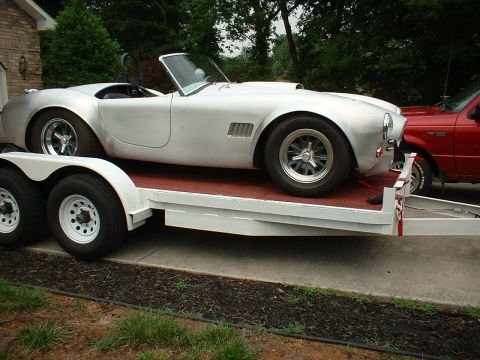 1965 Shelby Cobra - A silver 1965 Shelby Cobra is transported on a trailer, with a red pickup truck visible in the background.