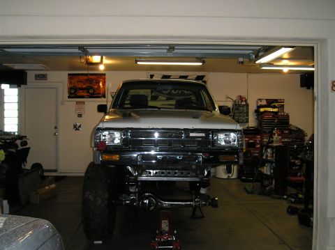 1986 Toyota Hilux - Head-on view of a lifted white Toyota pickup sitting on jack stands inside a home garage, front axle and steering linkage exposed below the…