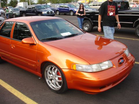 1993 Ford Taurus - An orange sedan with aftermarket wheels is parked in a lot with other vehicles.