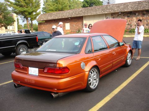 1993 Ford Taurus - This orange sedan features a contrasting brown trunk lid and aftermarket wheels, parked outdoors with the hood open.
