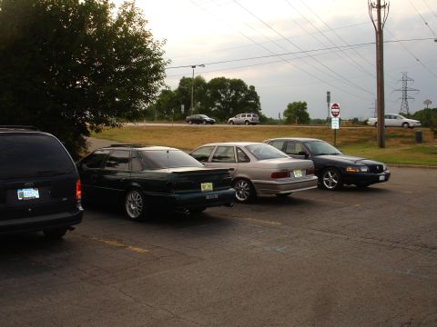 1993 Ford Taurus - Three sedans are parked in a lot under an overcast sky.