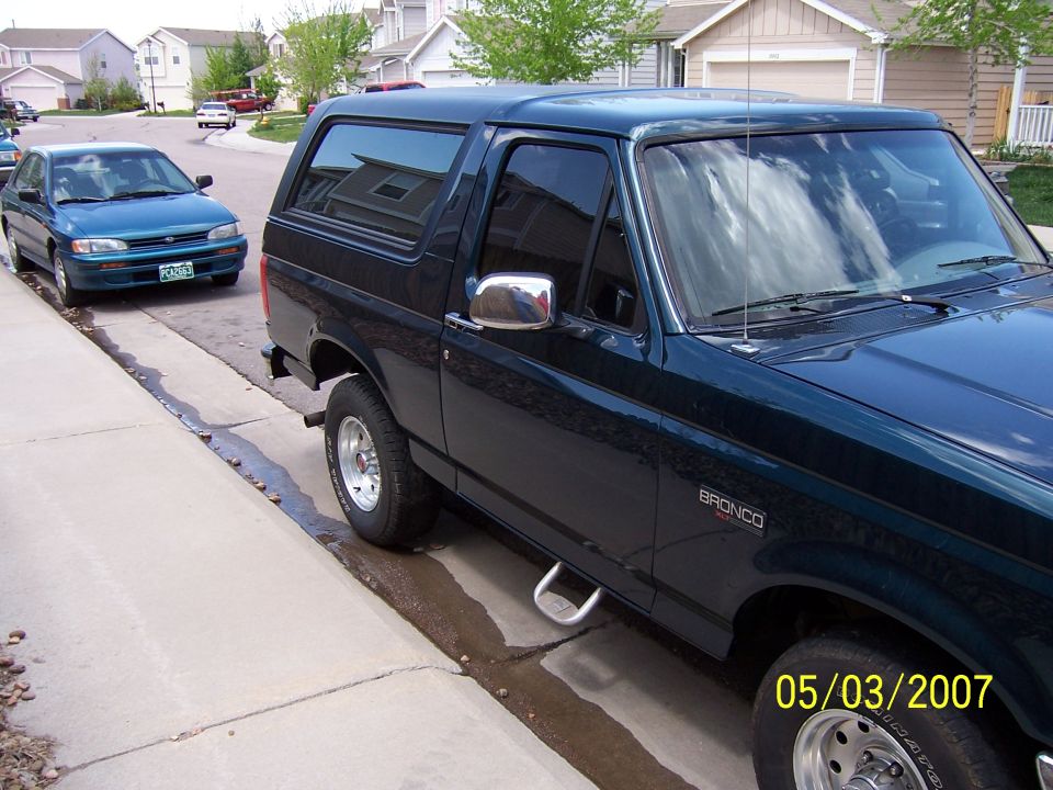 Dark green Ford Bronco SUV parked curbside, passenger side view, with a teal sedan behind it
