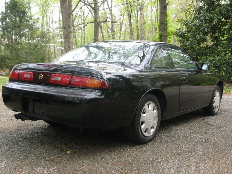 Black 1995 Nissan 240SX coupe, rear 3/4 view. The car is parked on a gravel surface with trees in the background
